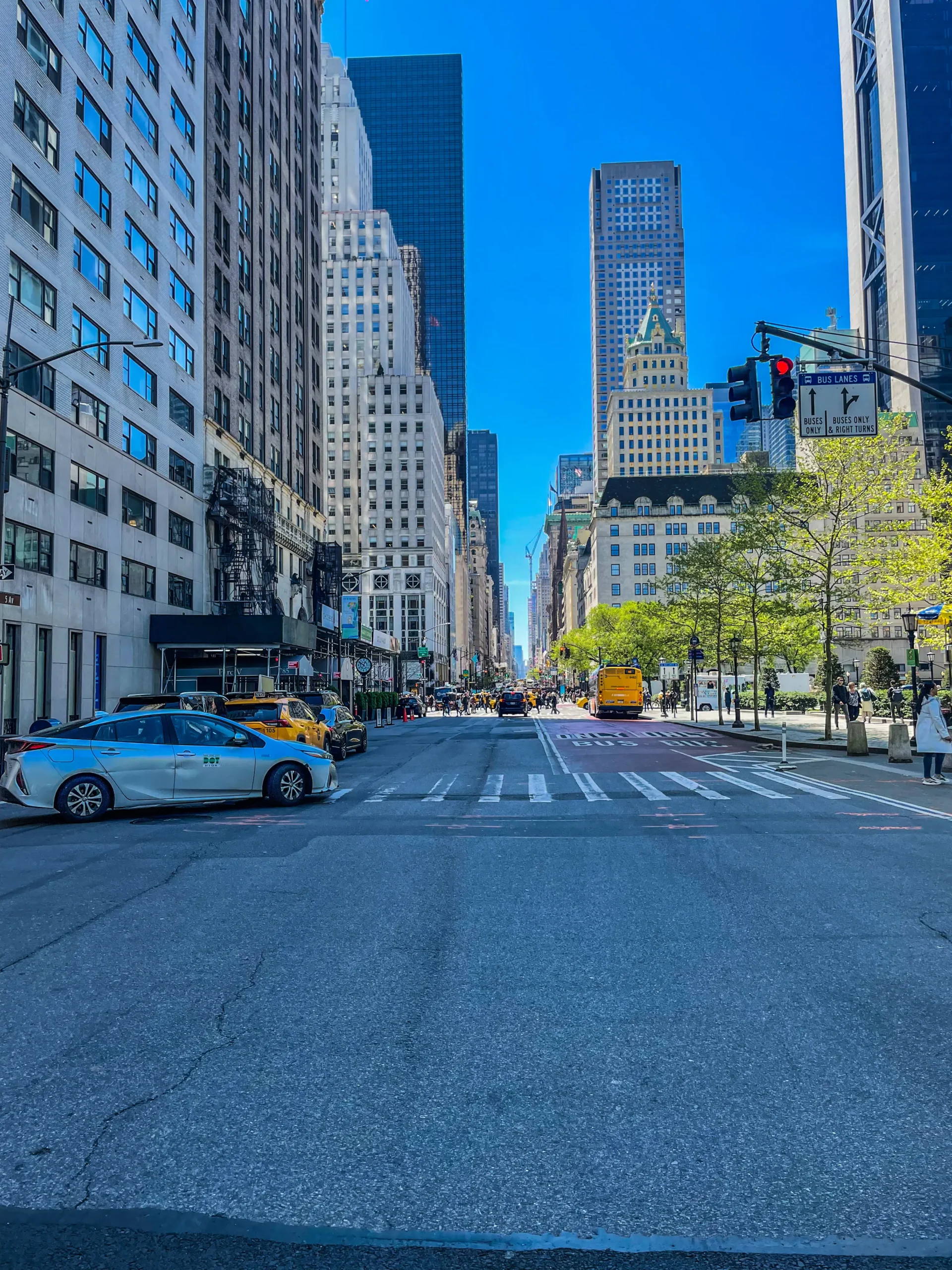 Low-angle view of a bustling New York City street with tall buildings lining both sides, taxis, and a clear blue sky. Midtown Manhattan skyscrapers are centered in the background.